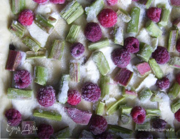 Раскатываем нужнное количество теста в пласт. Укладываем с небольшими бортиками на противень. Сливочное масло натираем на крупной терке и равномерно распределяем по тесту. Посыпаем поверхность сахаром. Сверху укладываем ревень и малину (в моем случае ревень и малина свежемороженые).