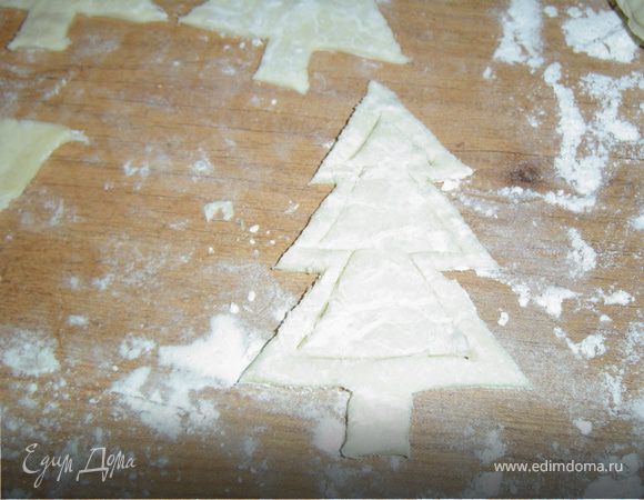 Раскатываем готовое слоеное тесто, вырезаем из плотной бумаги шаблон в виде елки, прикладываем его к тесту и вырезаем фигурки.