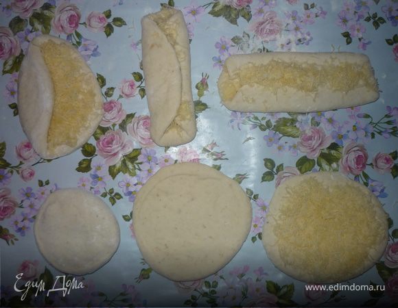 Вторую половину теста режем на 6 частей. Каждую часть раскатываем в круг толщиной 1 см. Посыпаем ровным слоем тертого на мелкой терке сыра, заворачиваем края к центру один на другой, слегка прикатываем вдоль скалкой.