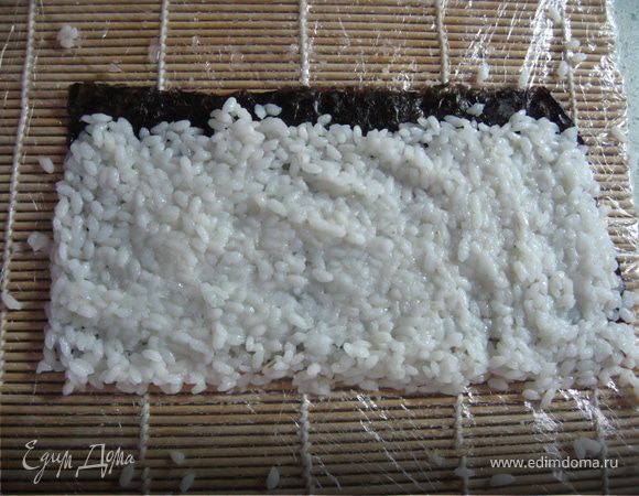 Приготовить коврик для заворачивания роллов,обернув его пленкой.Разрезать лист нори пополам,положить на коврик.Выложить на него рис как показано на фото,оставив 1-2 см нори свободным,а с другой стороны "заехать"за край.Чтобы рис не лип к рукам - смачивайте пальцы разведенной с уксусом водой