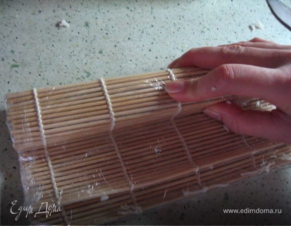 Поднимая ближний от себя край коврика и прижимая аккуратно вниз свернуть ролл.Так,что бы рис был равномерно распределен.Прижать пальцами коврик,чтобы получилась квадратная форма