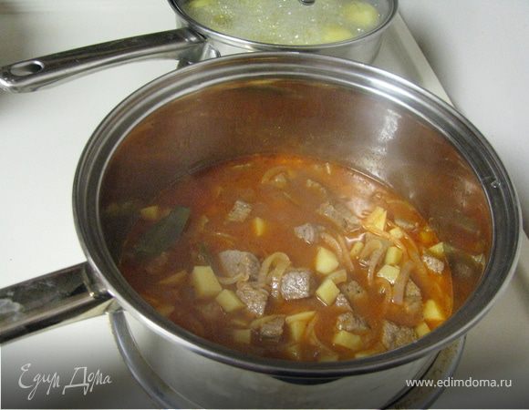 В той же сковороде слегка обжарить лук и картофель, снова виложить мясо, приправить пастой, паприкой и тмином. Влить бульон, довести до кипения накрыть крышкой и тушить на маленьком огне около часа периодически помешивая.