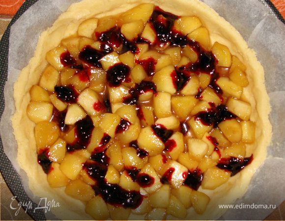 Нагреть духовку на 180-190 градусов, форму застелить пергаментом и смазать маслом. Тесто раскатать в пласт толщиной около 1 см, наколоть по всей поверхности вилкой, выложить в форму и сформировать бортики. На корж выложить яблочную начинку, добавить немного варенья или вишни. 