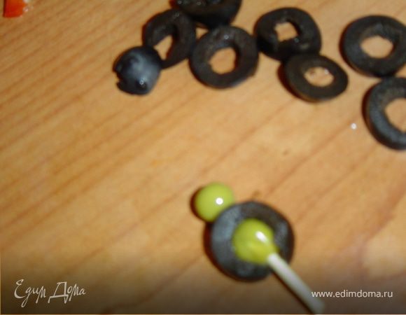 Нарежьте маслины поперек кружочками, кружочки нанизывайте на зубочистки, насадив горошины внутрь и сверху.