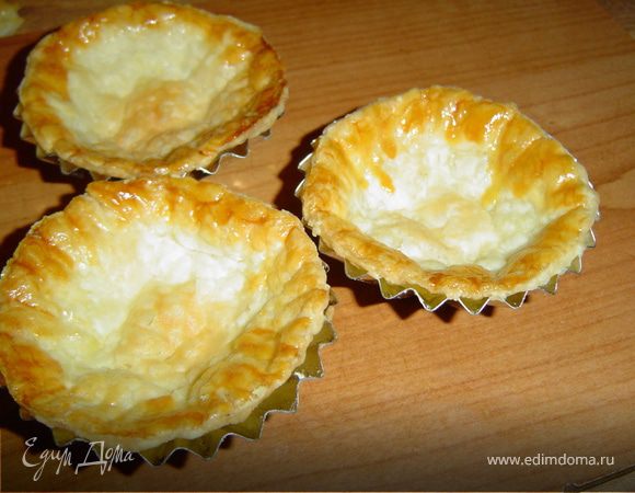Раскатайте готовое слоеное тесто, вырежте стаканом кружочки и распределите их по формам, смазанным маслом. Края тарталеток смажьте желтком. Выпекайте при температуре 180°С , до золотистого цвета.