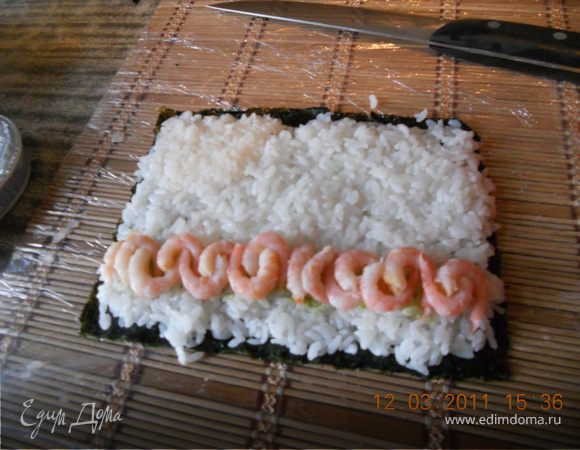 Лист нори разрезать пополам(ровно посередине)уложить на коврик(коврик предварительно обернуть пищевой пленкой,тогда рис не будет прилипать)
на водоросль выложить рис,равномерно распределить.Чтобы рис не прилипал к рукам смочите их в уксусе.
Зытем на рис выкладываем сыр,креветки,брусок авокадо,брусок огурца,рыбку и аккуратненько заворачиваем.