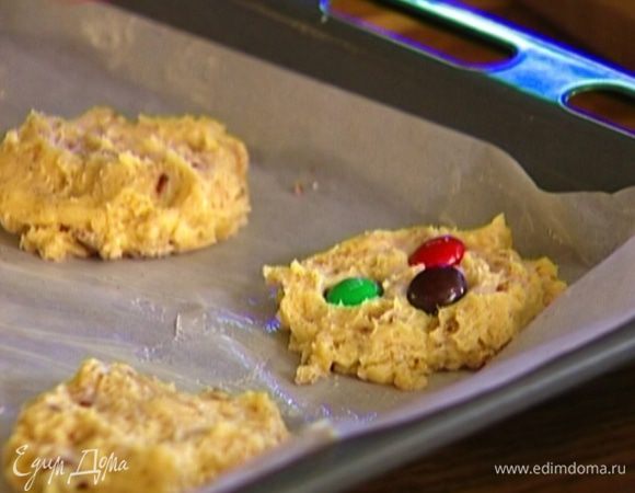 Из теста сформировать шарики, выложить их на противень, слегка прижать руками и украсить печенье конфетами, немного вдавливая их в тесто.