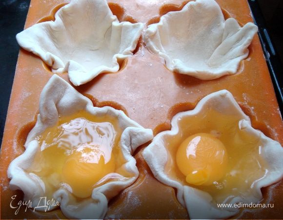 С вечера переложить слоёное тесто из морозильника в холодильник.Утром разогрейте духовку до 180*С, раскатайте тесто и разрежьте его на квадраты по размеру ваших формочек.У меня вышло 10 штук.Выложите квадраты в смазаныё маслом формы(силиконовые не смазывать),немного придавить их руками и выпустить в каждую тарталетку по одному яйцу.