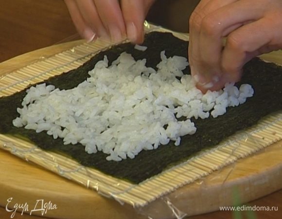Застелить коврик для суши пищевой пленкой, положить сверху лист нори и выложить на него слой суши-риса толщиной примерно 9 мм. 