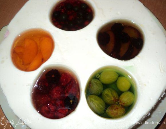 Вынуть торт из холодильника. В стаканы по очереди налить немного горячей воды, чтобы было легче вынуть их из формы. В образовавшиеся выемки положить по  немного ягод и фруктов,  залить охлажденным желе соответствующего цвета и поставить в холодильник для застывания на 2 часа ( немного желе оставить ).