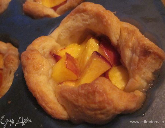 Слоёное тесто немного раскатать, разрезать на квадраты 10х10. Придавая форму корзиночек,  уложить в тефлоновые формочки для кексов.
На дно каждой корзиночки положить порезанные мелким кубиком персики и насыпать чайную ложку сахара. Запечь в предварительно разогретой до 180 градусов духовке минут 15. Остудить. Украсить взбитыми с сахарной пудрой сливками и ягодами. Приятного чаепития!