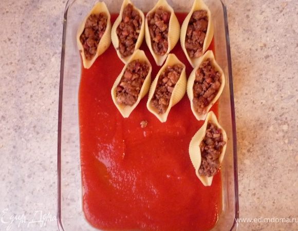 Разогреть духовку до 180°C. Форму для запекания смазать маслом. Оставшееся томатное пюре посолить, поперчить, добавить чили и щепотку сахара и выложить в форму. Ракушки нафаршировать мясом
и также выложить в форму.