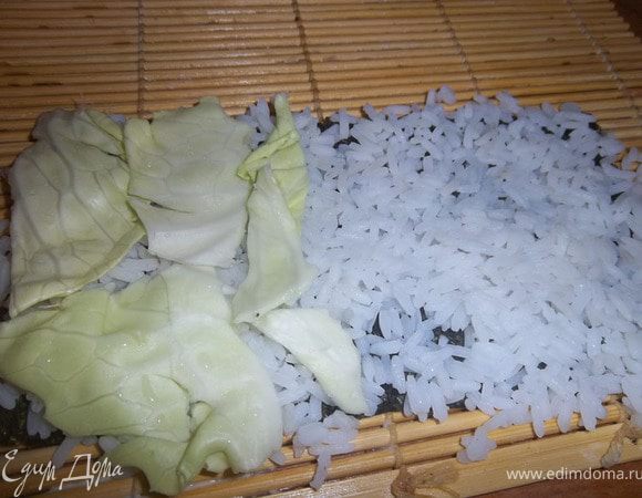 Положить на соломенный коврик лист нори. Сверху - отварной рис. Разровнять, слегка уплотнить. На ½ нории выложить лист капусты. Оставить вторую половину листа нори свободным.
