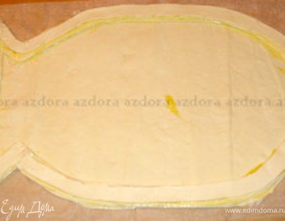Переложить вырезанную рыбку на противень вместе с бумагой для выпечки. Взять второй пласт слоёного теста. Тесто нарезать на полоски шириной около 1,5- 2 см.
Из этих полосок  сделать бортики, чтобы удержать начинку. Для этого взбить яйцо, смазать края вырезанной рыбки и приклеить полоски теста по всему периметру рыбы. Сделать три слоя из полосок. Из остатков этого пласта вырезать хвост и голову рыбки.

