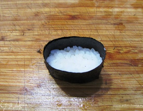 Заверните полоску вокруг риса по длинной стороне. Смочите край нори водой и сожмите между собой слои нори. Таким образом они слипнутся.
Поставьте получившийся "боевой корабль" на стол. Слегка прижмите сверху рис, чтобы он полностью распределился внизу. У вас получится некая лодочка с бортами из нори и дном из риса.