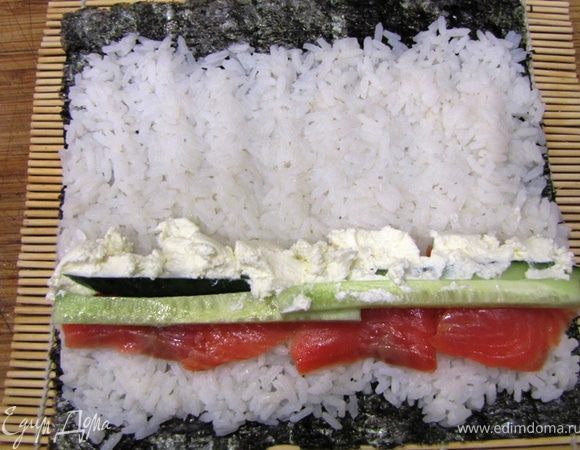 Теперь готовим Маки, то есть роллы
Я готовлю толстые роллы, поэтому использую целый лист Нори и довольно много риса.
Положите лист Нори на бамбуковый коврик.
Распределите сверху рис так, чтобы он полностью закрывал поверхность листа нори, но не доходил три-четыре сантиметра до дальнего конца.
Учтите, чем ровнее вы уложите рис (особенно это касается боковых краев листа нори), тем плотнее и аккуратнее у вас получится ролл.
Выложите полоски рыбы, огурца как показано на фотографии. Рекомендую положить по две-три полоски. Если у вас есть шприц, выдавите с его помощью полоску сливочного сыра. Толщина полоски должна быть аналогична толщине рыбы (т.е. если три полоски рыбы, то и толщина полоски сыра должна быть как у трех полосок рыбы).