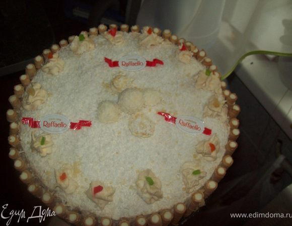 Нанести поверх ягод тонким слоем крем.
 15 конфет измельчить в блендере (охлаждённые конфеты измельчать намного удобнее) и посыпать поверх крема.Накрыть вторым коржом.Оставшимся кремом смазать верх и бока торта.Украсить конфетами „Raffaello“ выкладывать по желанию
 В заключении, обсыпать торт со всех сторон кокосовой стружкой.Я ещё украсила взбитыми сливками, цукатами и трубочками!