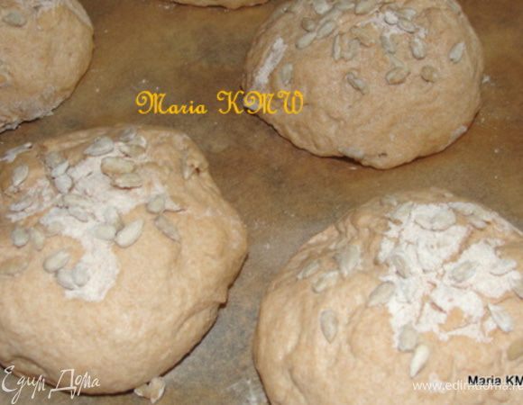 Духовку предварительно разогреем до 200 градусов. 
Тесто обминаем и формируем из него 12 круглых булочек.
Выкладываем их на противень, с бумагой для выпечки и даем немного подойти.
После того, как булочки увеличились, надрезаем верх крестом, смазываем водичкой и посыпаем оставшимися семечками.