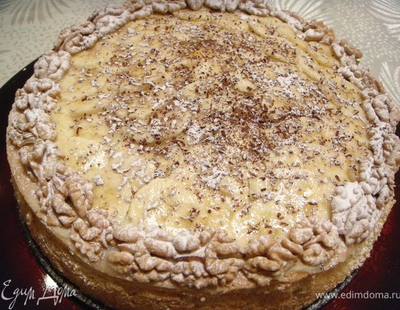 Накрыть сверху срезанной макушечкой. Смазать оставшимся кремом. Сверху выложить порезанный тонкими кружочками банан. По периметру выложить грецкие орехи и посыпть сахарной пудрой и тертым шоколадом.