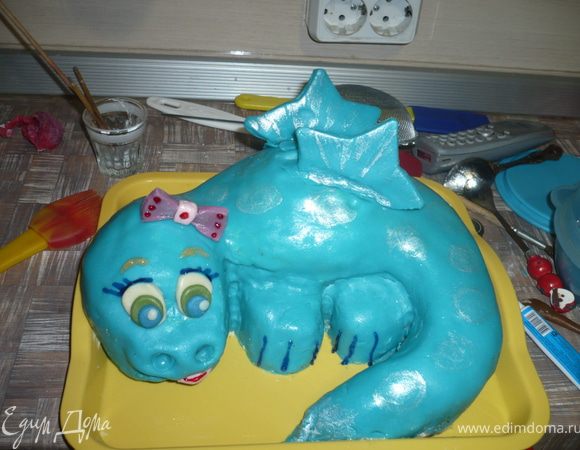 Украсить нашего дракончика - выдавить ноздри, глазки сделать из овальчиков белой, зеленой и синей мастики, вылепить бантик, сделать язычок из розовой мастики, прилепить крылья -  я вырезала из из большой формочки для печенья - звезды, потом я пробежалась серебристым кандурином по дракончику, гелевыми пищевыми фломастерами нарисовала ресницы, брови и кое-где нанесла последние штрихи. Торт-Дракон готов!