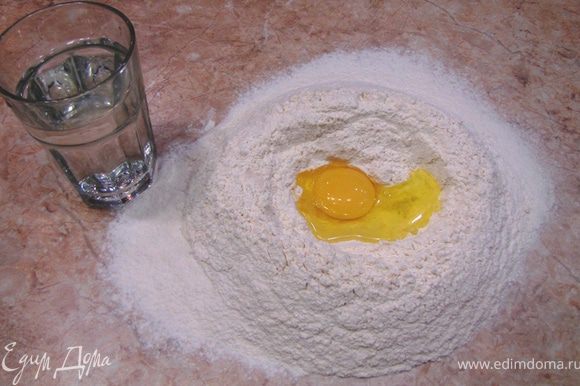 Для начала приготовим лапшу. Просейте через сито муку на стол или большую доску. Сделайте посередине ямку. Разбейте туда яйцо и вылейте воду. Аккуратно вилкой вмешивайте изнутри муку в жидкость. Затем замесите тесто. Тесто должно получиться крутым.