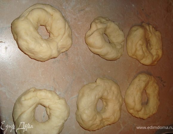Затем разделить тесто на 6 равных частей, не подпыляя стол мукой, подкатать в тугие шарики. Слегка расплющить шарик, указательный палец окунуть в муку и проткнуть им шарик посередине. Постепенно увеличивать отверстие, вращая тесто. Оставить бублики на 5-10 мин., прикрыв влажным полотенцем, затем растянуть до нужного размера.