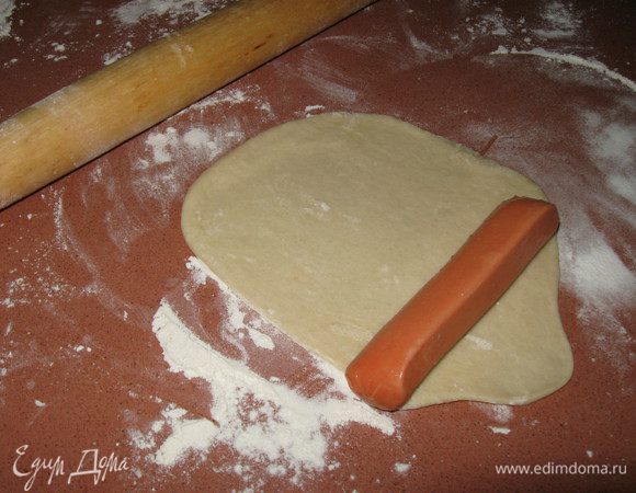 Готовое тесто на дрожжах разделите на шарики. Каждый шарик раскатайте скалкой, положите на край сосиску.