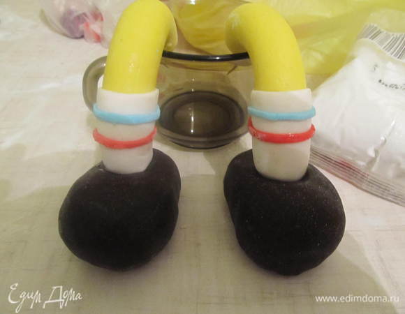 Каждую ножку одеть в беленький носочек и прекрепить по 2 полосочки (голубая, красная). Одну сторону ножек, с помощью зубочисток настромить на туфельки, другую сторону к штанам.