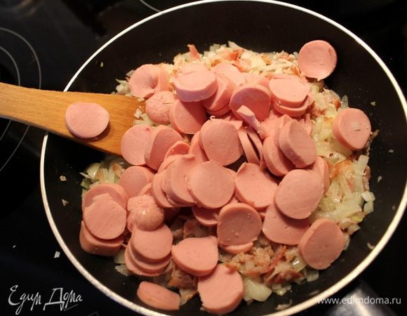 Добавьте к луку с беконом нарезанные тонкими кружками сосиски. Обжарьте все вместе несколько минут. Выложите смесь на отварное мясо в горшок.