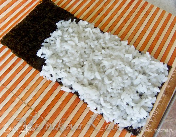 Лист нори разрезать пополам. Выложить на лист немного риса, разровняв его ровным слоем, оставив немного пустого места на листе. 