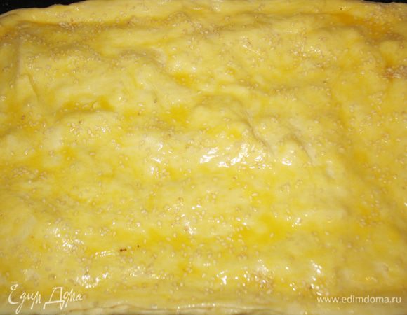 Поверх теста выложить начинку. Желток смешать с молоком. Края теста смазать яичной смесью. Накрыть начинку оставшимся тестом и защипнуть края. Оставшейся яичной смесью смазать тесто и посыпать его кунжутом. 