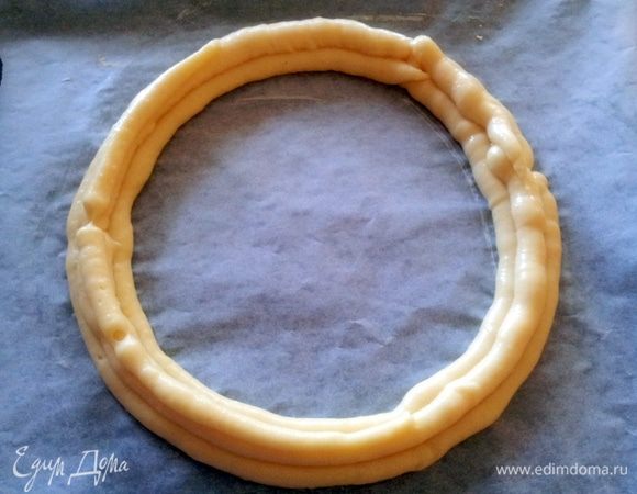 Приготовить противень, застеленный пергаментом и на пергаменте  нарисовать круг диаметром 24-26 см. Выдавить из кондитерского мешочка заварное тесто в виде кольца. Затем второе кольцо выдавить внутри первого, А третье разместить между первыми  и вторым. 