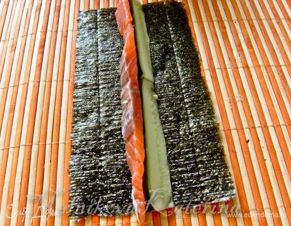 Лист нори разрезать пополам, выложить на циновку. На лист выложить огурец (примерно посередине), за ним выложить полоску лосося. 