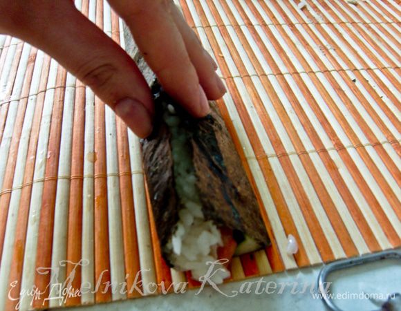 Пальцы смочить в растворе специального уксуса для суши и воды (они должны быть влажными). Край, ближний к огурцу, загнуть, формируя пальцами что-то вроде сектора. 