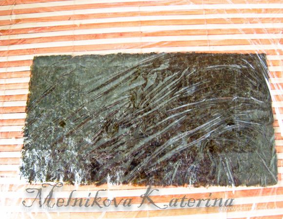 Для формирования такого ролла нужно иметь под рукой пищевую пленку, которой оборачивается циновка для суши. Я буду показывать с использованием половинки листа нори, который выступает в качестве шаблона для накладывания риса, но можно обойтись и без него, просто выложить рис ровно в виде небольшого прямоугольника. Итак, сначала нужно положить на циновку половинку листа нори, накрыть пищевой пленкой, закрепить ее за край циновки.