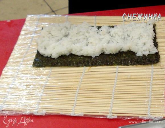 На нижний край коврика кладем нори, смоченными руками распределяем рис по поверхности нори. Делайте это уверенно, легкими движениями, двигая от центра к краю. Верхний от Вас край нори оставляем свободным!