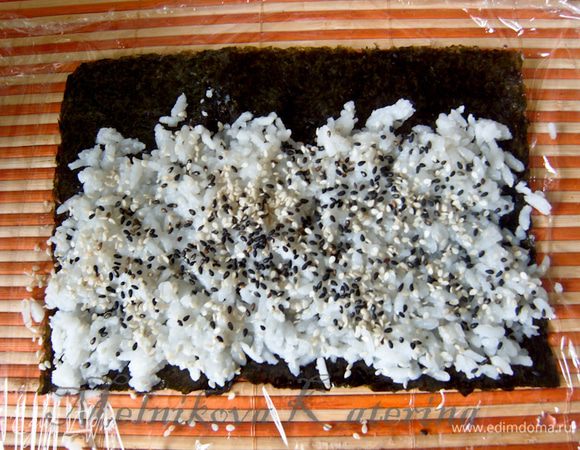 Для приготовления суши-ролла с рисом на изнанку необходимо сперва наложить на коврик для суши пищевую пленку, оставив внизу кусок длиной примерно 15 см. На пленку выложить половину листа нори. На нори выложить рис, оставив пустой полосу с одного края длиной шириной примерно 1.5 см. Рис разровнять. Присыпать сверху кунжутом. 