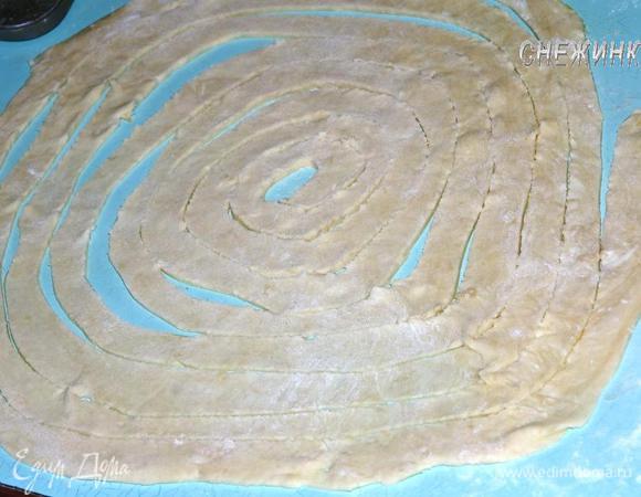Слоеное тесто раскатываем в круг, постоянно присыпая мукой. Вырезаем одной сплошной полоской. Начинаем от края и, двигаясь по кругу, надрезаем полоску одинаковой ширины.