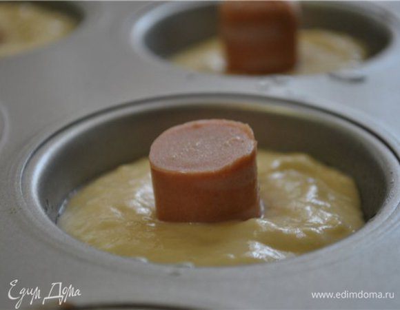 Смешать венчиком все жидкие составляющие.
Муку, соль и разрыхлитель смешать в отдельной миске.
Соединить и хорошо размешать.
Каждую сосиску разрезать на 3-4 части (в зависимости от длины сосиски).
Формы для кексов смазать маслом, наполнить на 2/3 тестом, в центр вставить сосиску. 