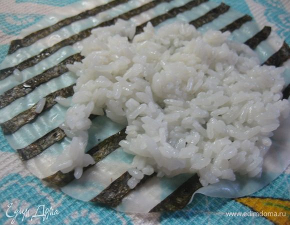 Лист рисовой бумаги опустить в теплую воду и дать ему размокнуть, он должен стать эластичным (у меня заняло около 30 секунд +/-).
На матису положите чистое сухое полотенце, на него выложите размокший лист рисовой бумаги и разложите на нем полоски нори. Т.к. у меня бумага оказалась круглая, то мне пришлось подрезать полоски нори, но если бумага будет прямоугольной, то полоски будут как раз по размеру.
Сверху распределите рис, смачивая руки в холодной воде, подкисленной уксусом.