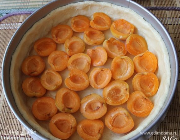 Разъёмную форму застелить пергаментом, бока смазать маслом. Тесто немного раскатать, перенести в форму и распределить по дну и стенкам, обмакивая пальцы в муку (так как тесто очень мягкое и немного липкое). Выложить половинки абрикосов по дну кожурой вниз.