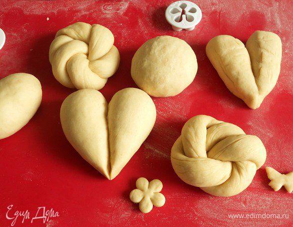 На рабочей поверхности разделите его на 12 равных кусочков примерно по 70 г, сформируйте их них любые по форме булочки (круглые, овальные, жгуты, сердечки). Для сердечек разрезать кусочек теста на 2 части, скатать 2 конуса, сложить острыми концами вместе. Соприкасающиеся поверхности слегка смочить водой. Сверху можно украсить мелкими фигурками из теста. Для витых булочек раскатать тесто в колбаску длиной 40 см, свернуть пополам в жгут, а затем его в кольцо, спрятав кончики внизу.