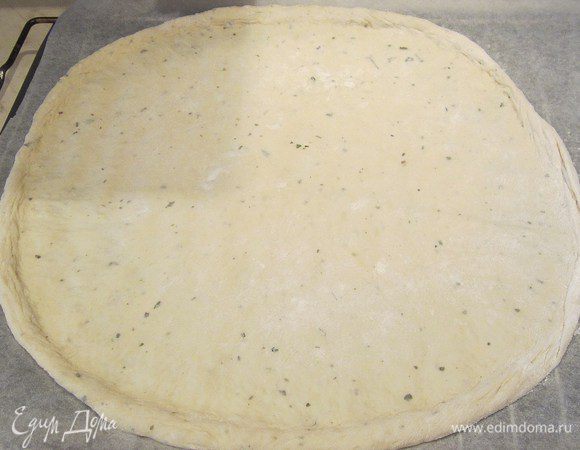 Вынуть готовое тесто и раскатать в круг на бумаге для выпечки, посыпанной мукой, стараться раскатать тесто тонко, сделав бортики по краям.А вообще.по-опыту,лучше тесто  не раскатывать,а руками сформировать круг,тесто не потеряет структуры пышности и будет намного вкуснее! Смазать кисточкой оливковым маслом.
