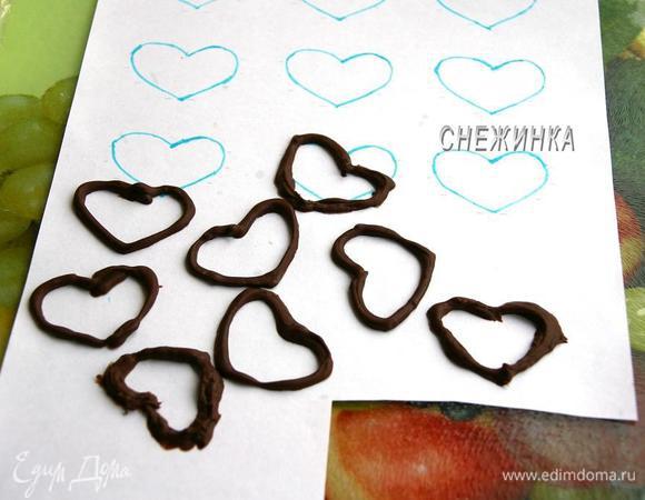 Подготовим украшения. Чтобы сделать сердечки из шоколада берём 40 г. шоколада, растапливаем его на водяной бане. Заготовим шаблоны сердечек. Я нарисовала сердечки на бумаге, положила лист на доску, а сверху накрыла пищевой плёнкой, хорошо закрепив края, чтобы плёнка не смещалась. Когда шоколад готов, перекладываем его в полиэтиленовый пакетик, отрезав кончик, и рисуем шоколадом по шаблону. Даём застыть в холоде. Сердечки легко отсоединяются.