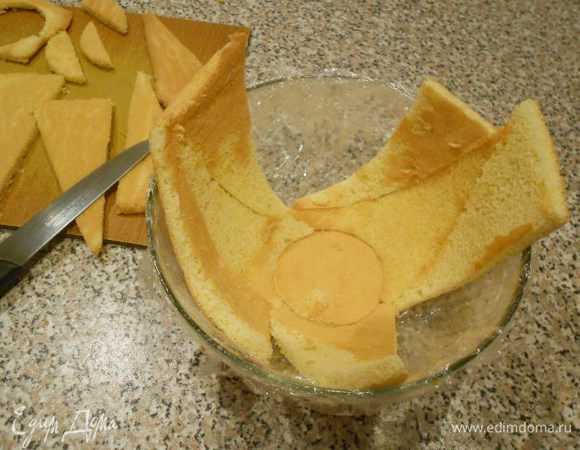 Из белых коржей вырезать тонким стаканом 1 маленький круг и один большой по диаметру миски. Остальной бисквит нарезать длинными треугольниками. Миску выстлать пищевой плёнкой. На дно положить маленький круг и плотно к нему укладывать треугольные кусочки.