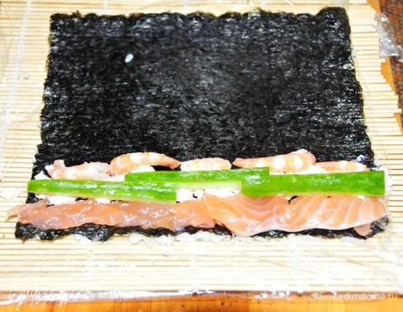 Третий ролл – Урамаки (ассорти):
На лист нории выкладываем рис. Потом аккуратно лист переворачиваем. Рис оказывается внизу, а нори сверху. На край выкладываем начинку: форель, полосочка сыра «Филадельфия», несколько креветок и полосочки огрурчика. Сворачиваем ролл.

