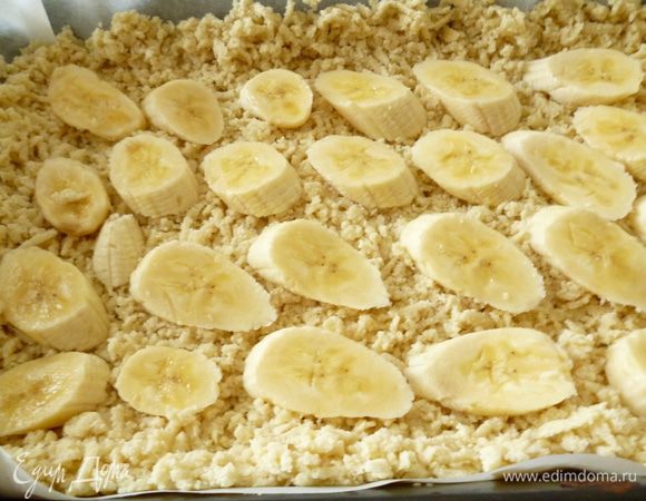 Форму для выпечки выстилаем пергаментом, смазываем маргарином и посыпаем манкой. 6 частей теста натираем через крупную терку, равномерно распределяем и формуем бортик. Выкладываем слой нарезанных бананов.
