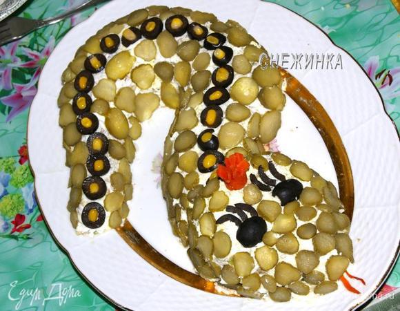 Теперь нарезаем тоненькими кружочками огурчики и маслины. Кружочками маслин выкладываем узор на спине змеи. В пустые серединки вставляем кукурузу. Всю остальную поверхность змеи покрываем «чешуёй» из колечков огурцов.