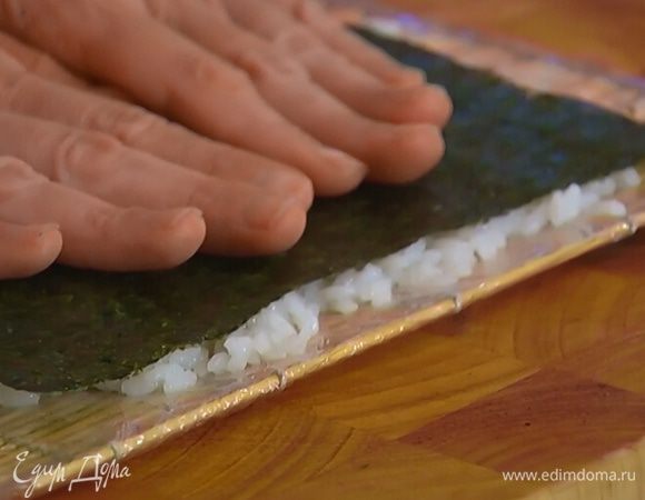 Коврик для суши обернуть несколько раз пищевой пленкой, выложить на него тонкий слой риса и утрамбовать его, сверху поместить лист нори ворсистой стороной к рису и прижать руками.