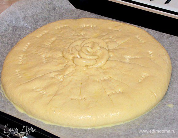 Стяните края теста к центру и закрепите, оставив небольшое отверстие. Пирог слегка прижмите, чтобы сделать его более плоским. Из оставшегося кусочка теста, сделайте лепестки и выложите их в центре в виде цветка.  Сделайте при помощи вилки несколько отверстий для выхода пара. Смажьте пирог смесью желтка с водой. Перенесите пирог вместе с пергаментом на противень и оставьте на 15 минут.